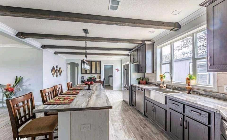 Spacious kitchen with rustic wood beams, a long island with wooden chairs, gray cabinets, and large bright windows. Warm, welcoming atmosphere.
