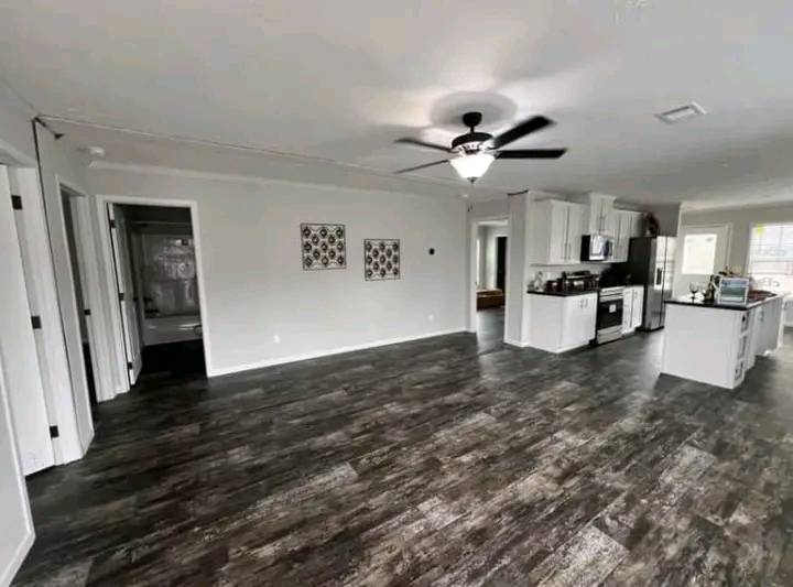 Spacious open-concept living area with dark wood flooring, white walls, ceiling fan, and a modern kitchen featuring white cabinets and stainless appliances.