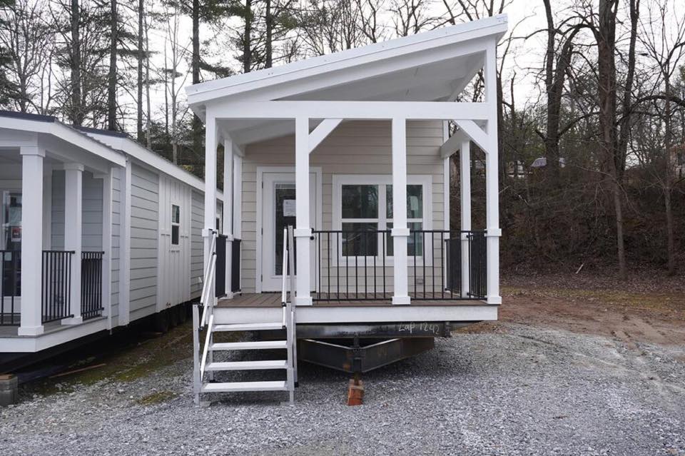 Small modern tiny house with a slanted roof, white trim, and front porch. Situated on gravel, surrounded by bare trees, creating a minimalist, rustic feel.