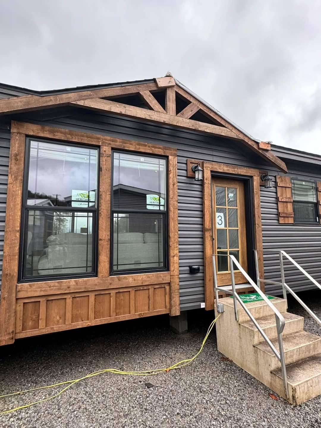 A modern tiny home with dark gray siding and wooden trim, featuring large windows, a wooden door marked with the number 3, and a small stone staircase.