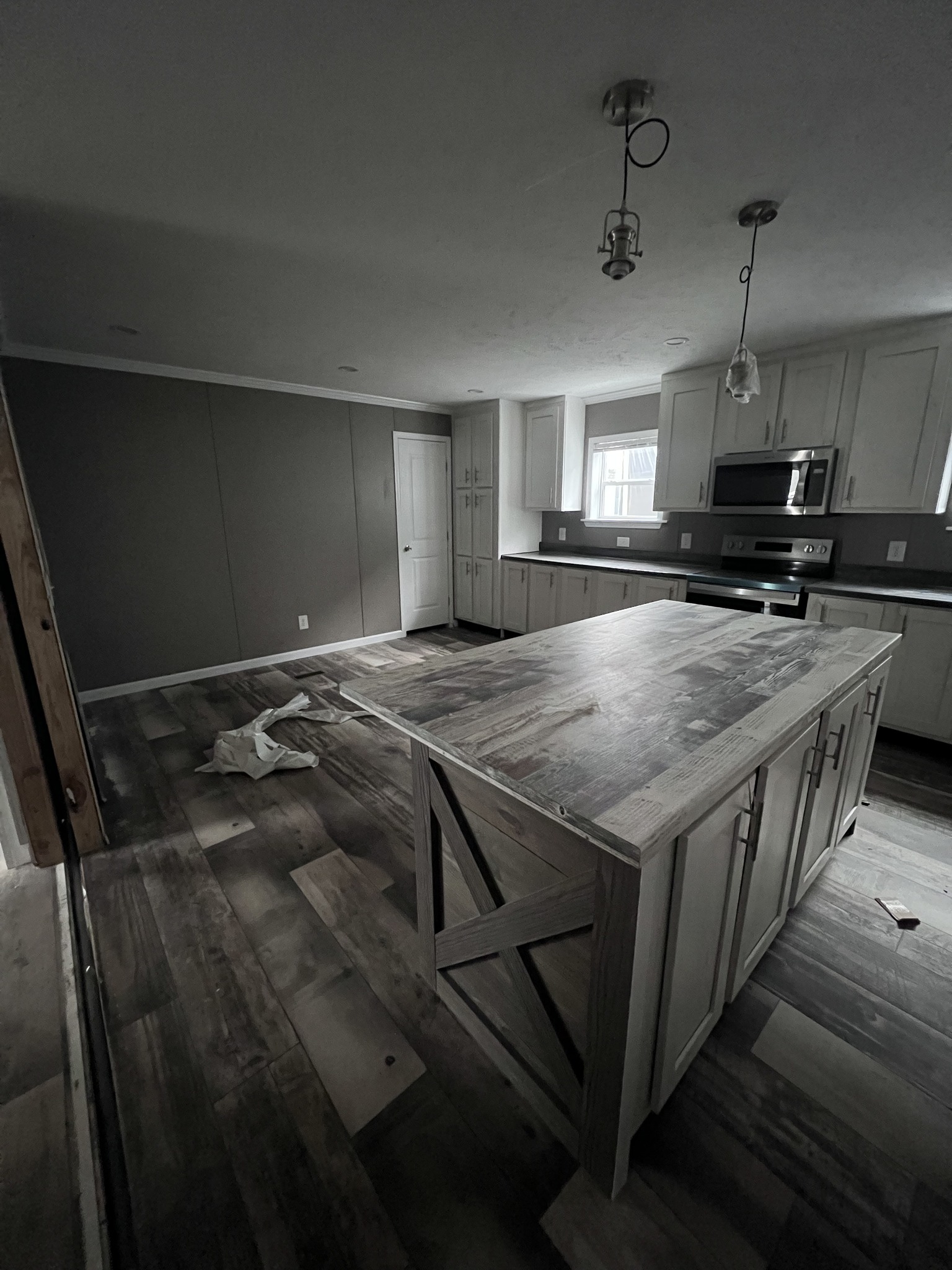 Dimly lit kitchen with rustic wooden island, white cabinets, stainless steel appliances, and dark countertops. A cloth lies on the wood floor.