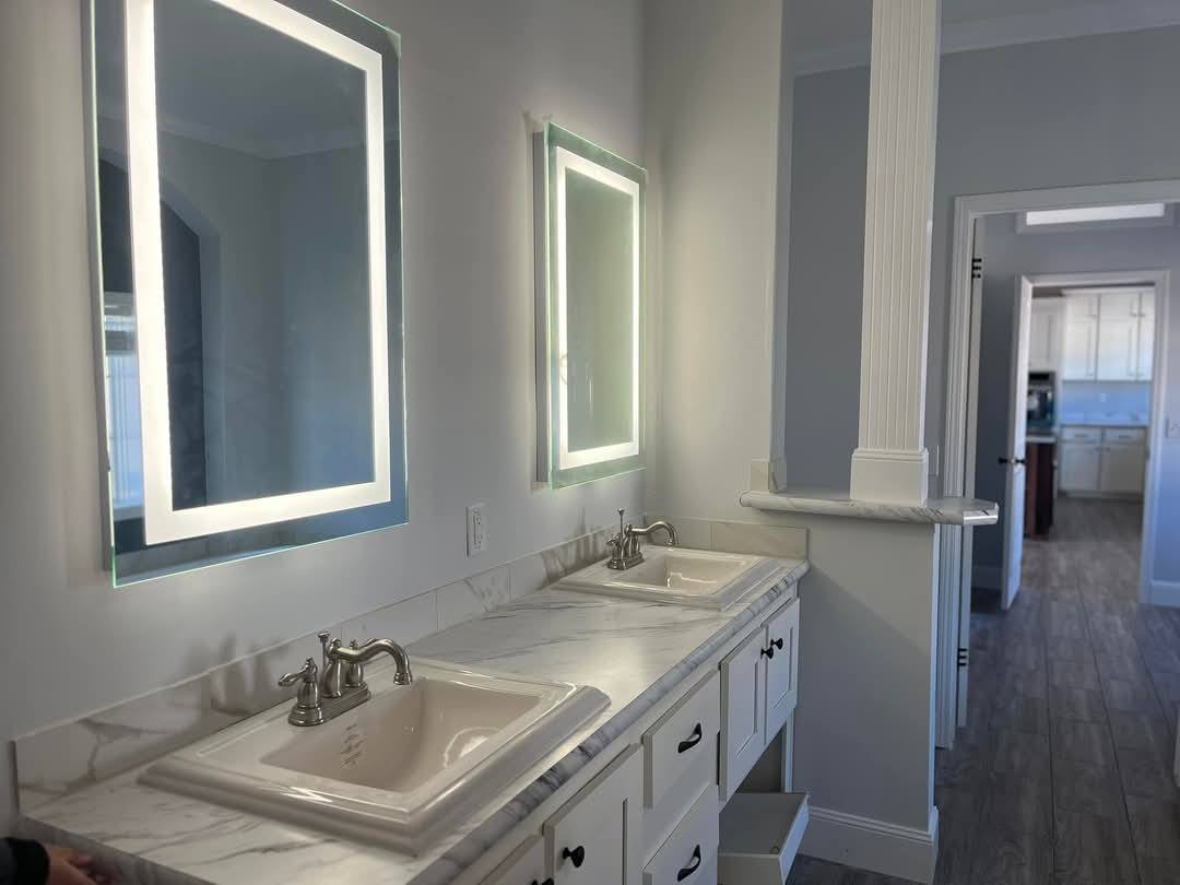Modern bathroom with dual sinks set in marble countertops, illuminated mirrors above, and a view into a kitchen with white cabinets and wood flooring.