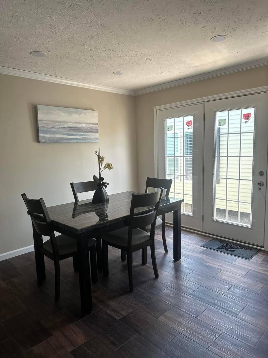 A cozy dining area with a dark wood table and four chairs on a polished wood floor. Light streams in through glass doors, creating a warm ambiance. A serene beach painting hangs on the beige wall.