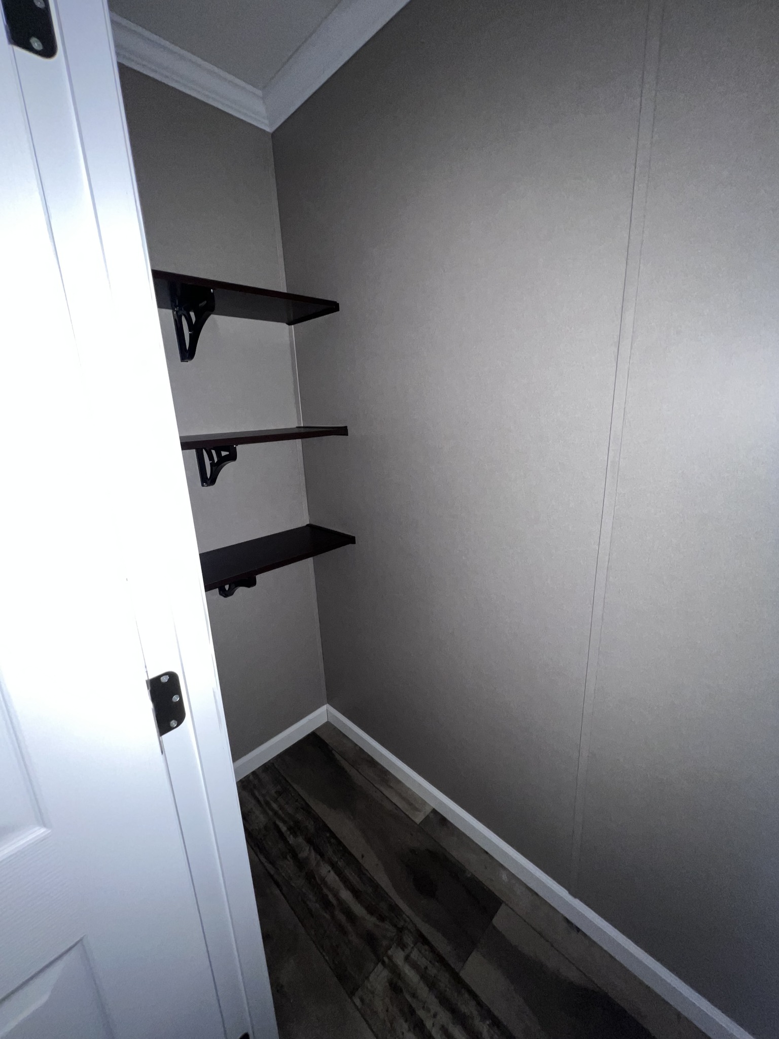 Small, empty closet with beige walls and two dark wooden shelves. The floor is wood-patterned, and a white door is partially open, revealing the space.