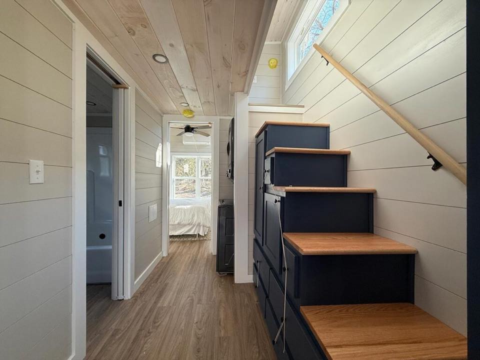 Narrow hallway in a tiny house with wooden floors and white shiplap walls. Compact stairs with storage lead to a loft, and there's a glimpse of a cozy bedroom.