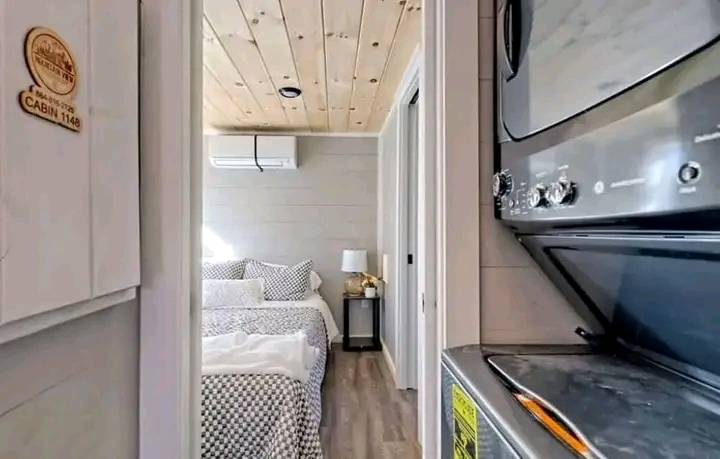 Cozy bedroom view from a hallway with a wooden ceiling and tiled floor. A neatly made bed is visible, next to a small nightstand with a lamp, conveying comfort.