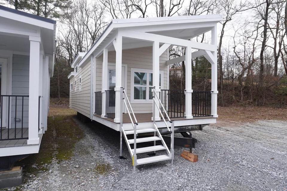 A tiny house with beige siding and white trim is elevated on stilts with a small porch. It has stairs leading to the entrance. The setting is a gravel lot with bare trees.