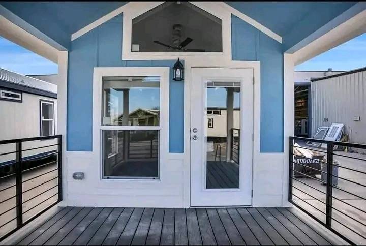 Cozy house with blue and white siding, featuring a glass front door and large window. The entrance has a wooden deck and black railing, under a clear sky.