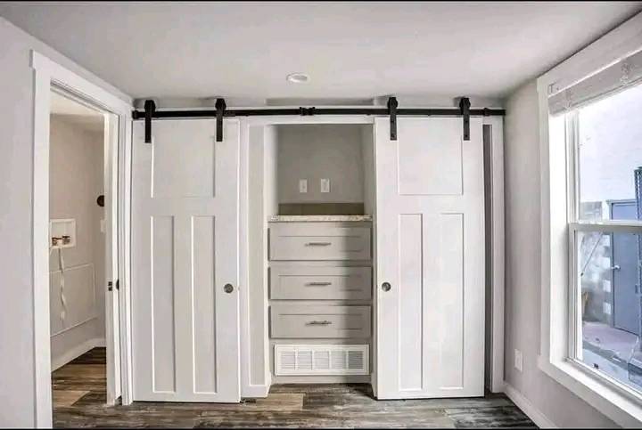 A small room with white walls features a closet with sliding barn doors, revealing stacked drawers. Adjacent are wood floors and a sunny window.