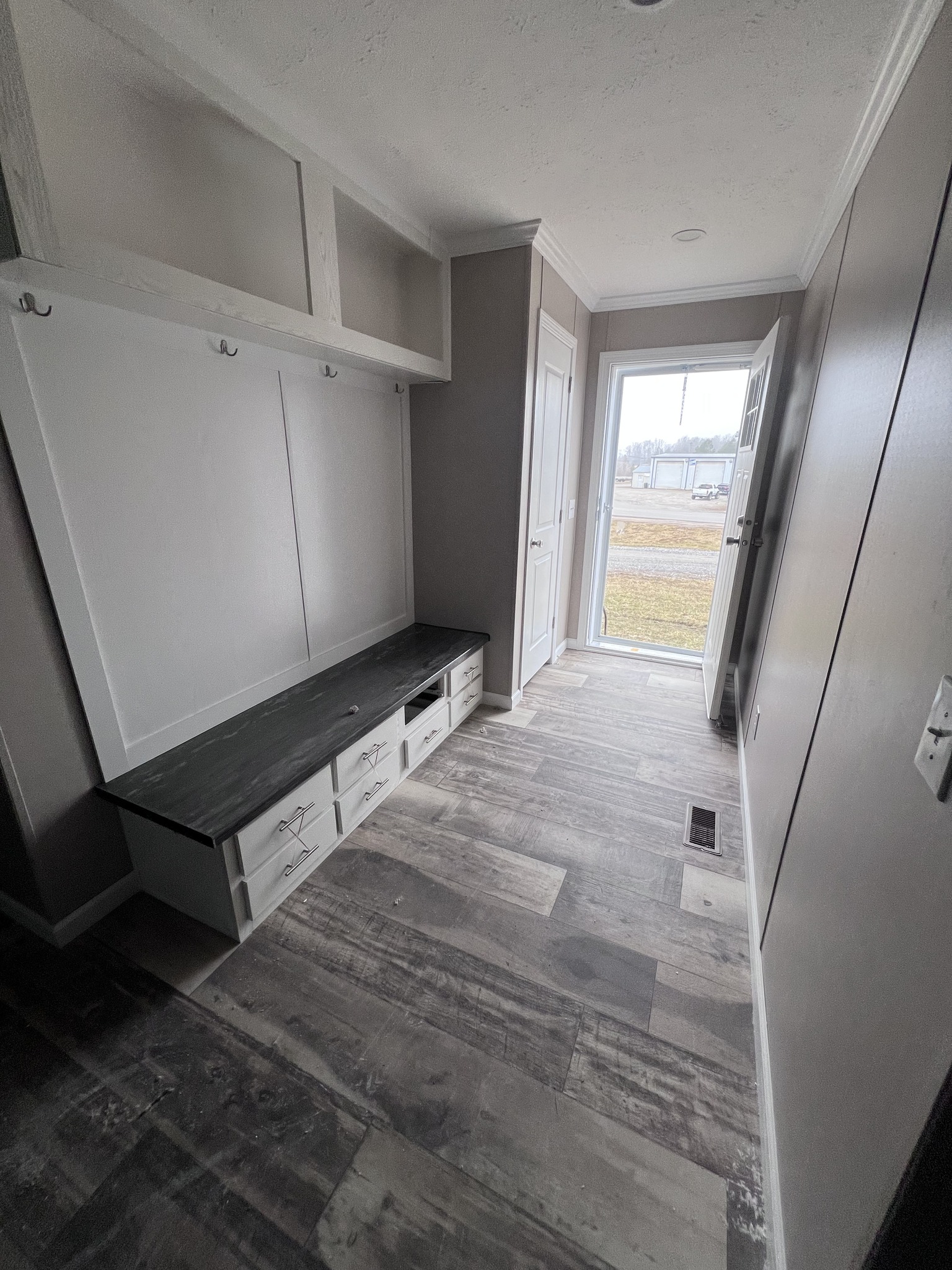 A narrow mudroom with light gray walls and wood-patterned flooring. It features a black bench with storage drawers and hooks above. A door with glass panels opens to a grassy yard, adding a sense of openness.