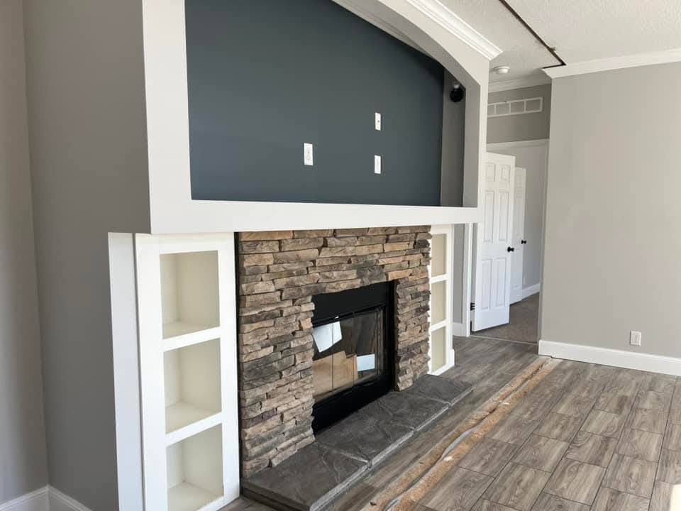 Living room with a rustic stone fireplace, white shelves on each side, and a gray accent wall above. Wood-style tile flooring adds warmth.