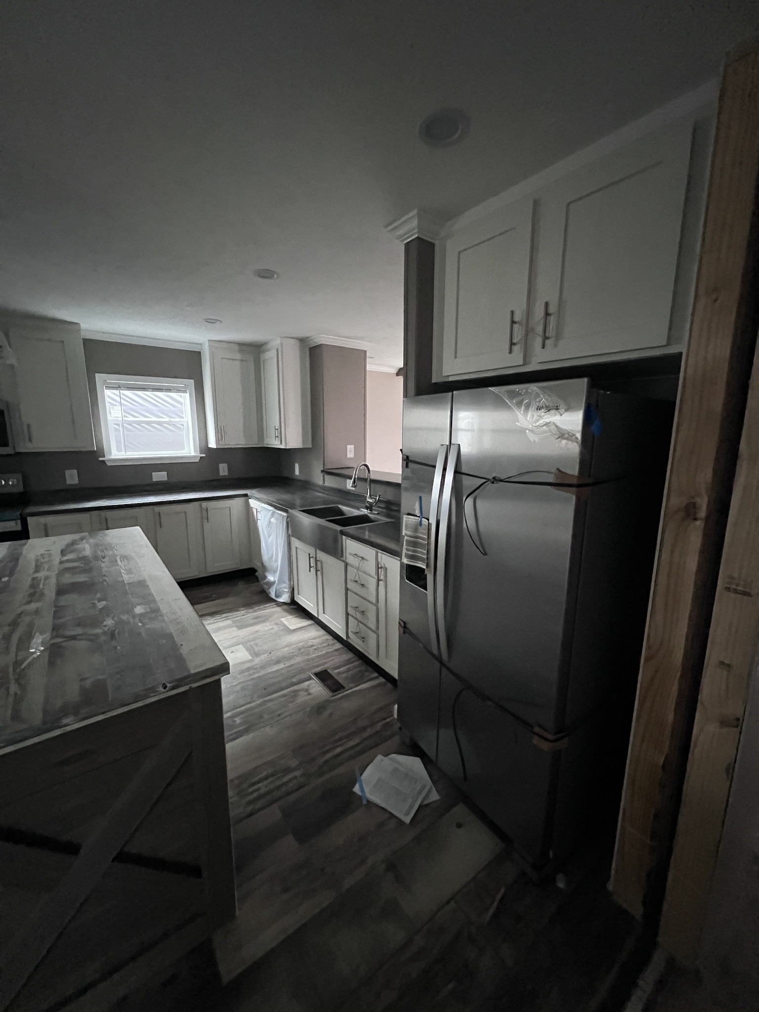 Dimly lit kitchen under renovation with white cabinets and a sleek stainless steel fridge. Wooden island and flooring add rustic charm.