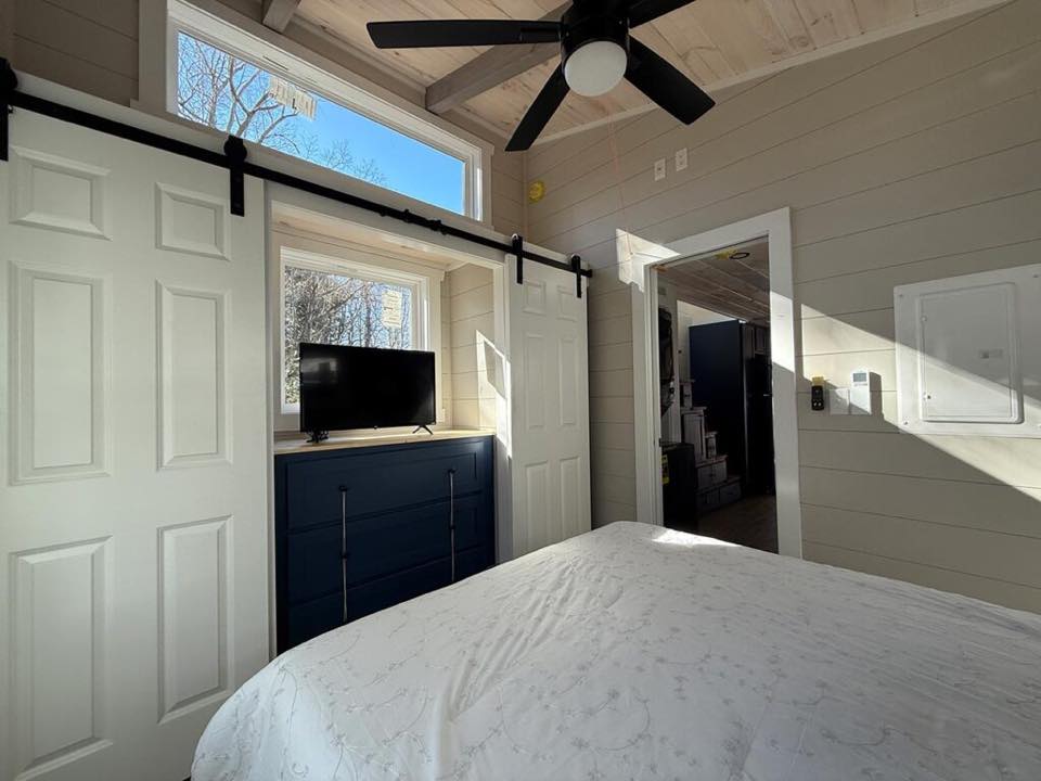 A cozy bedroom with a rustic feel featuring a white sliding barn door, ceiling fan, small TV on a blue dresser, and sunlight streaming through the window.