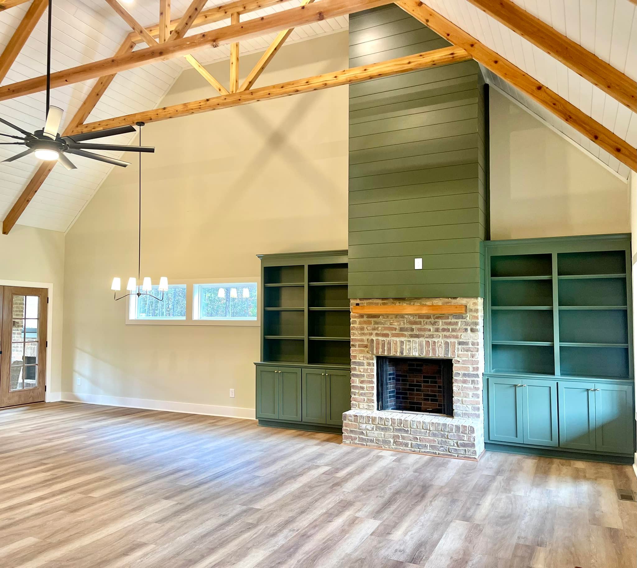 Spacious room with vaulted ceiling, wooden beams, brick fireplace, green built-in shelves, and large fan. Bright and airy with a modern rustic feel.