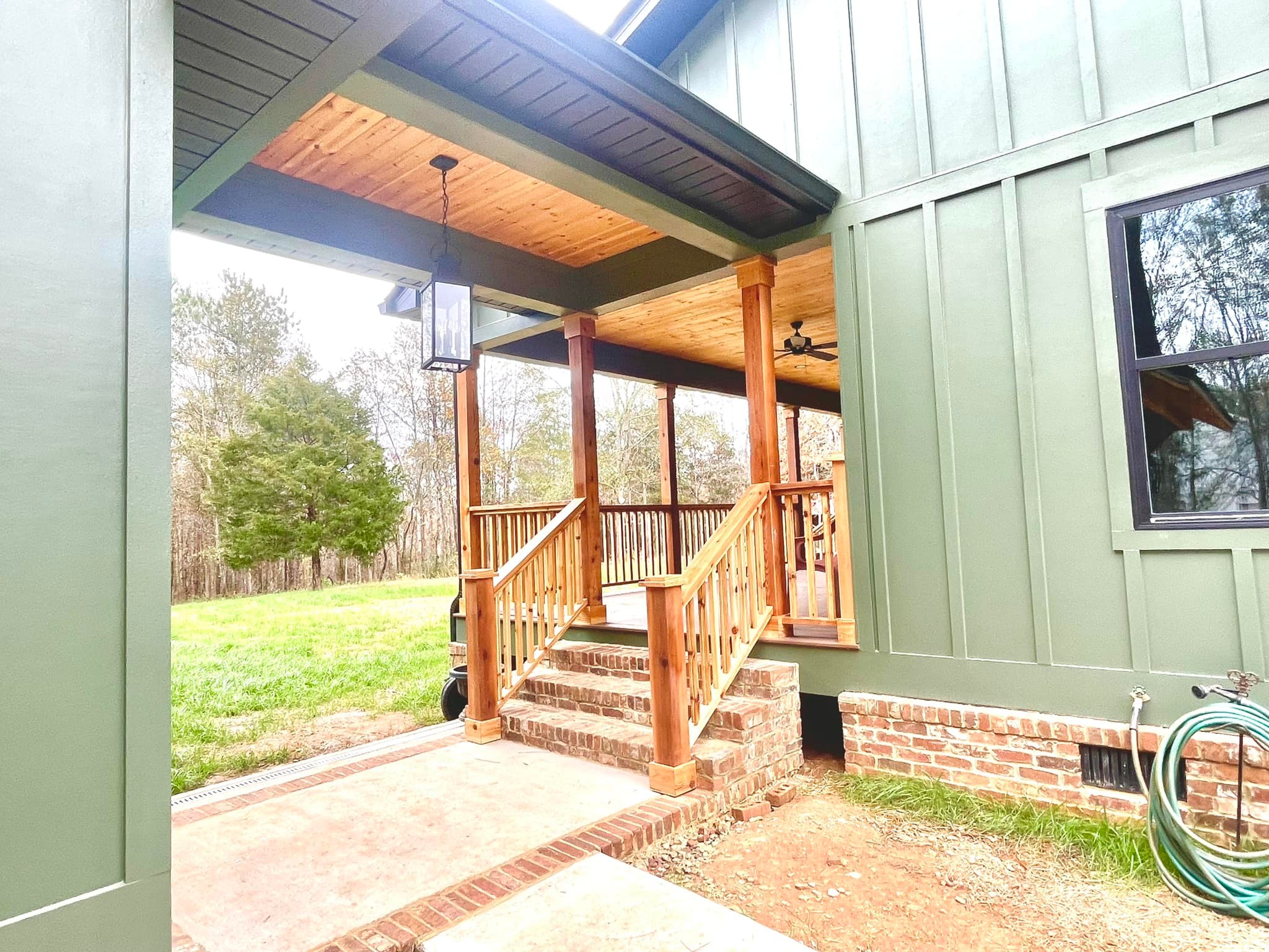 Wooden porch with a green exterior wall and gabled roof, brick steps and base. Surrounded by grass and trees, evoking a peaceful rural setting.