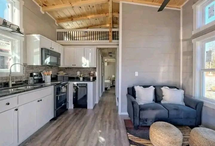 Modern tiny house interior with a light wood ceiling, gray walls, and laminate flooring. Features a compact kitchen, cozy gray sofa, and pouffe stools.
