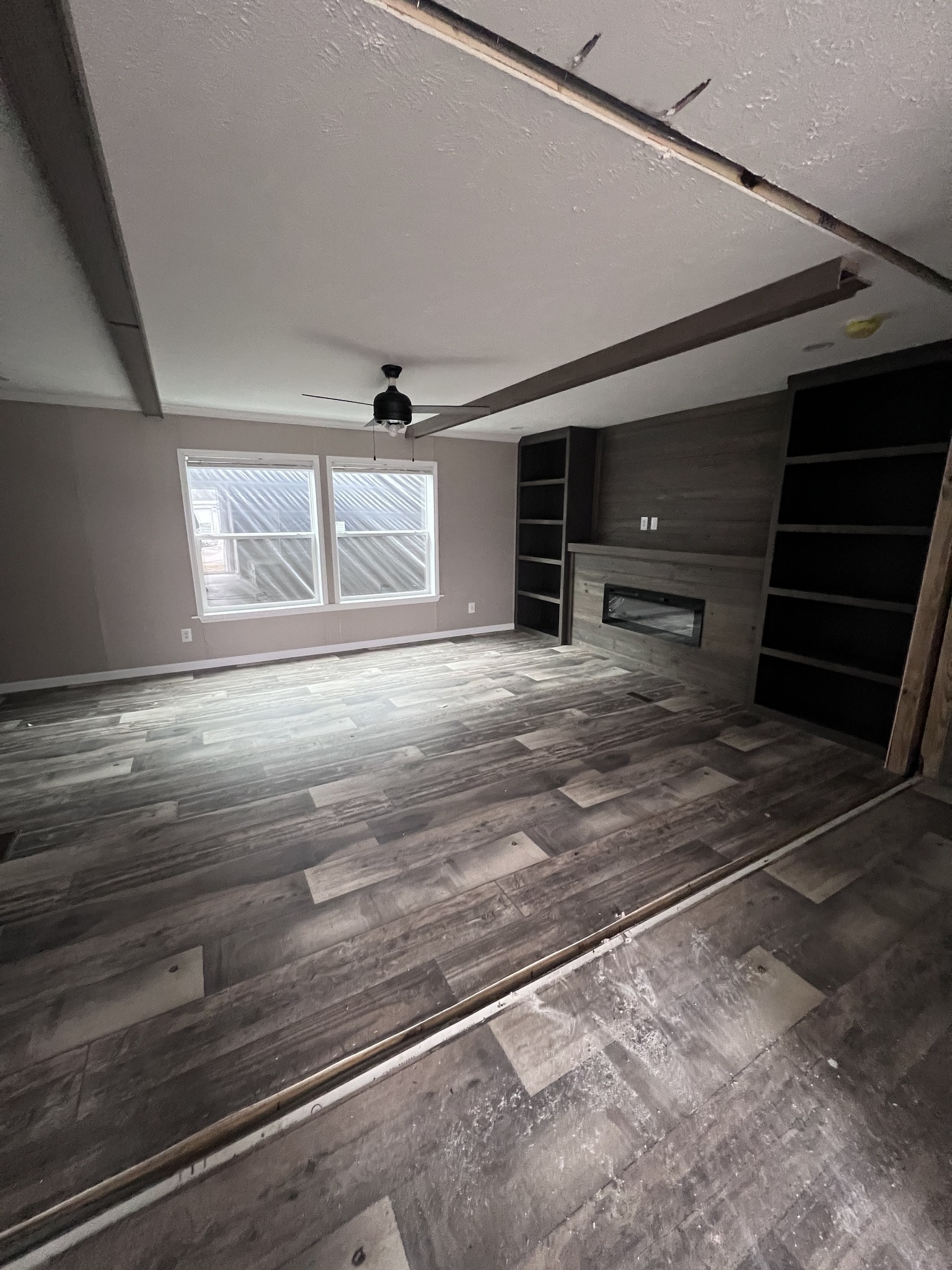 Spacious room with dark wood flooring, a ceiling fan, and large windows. Features a wall-mounted fireplace flanked by shelves, creating a modern ambiance.