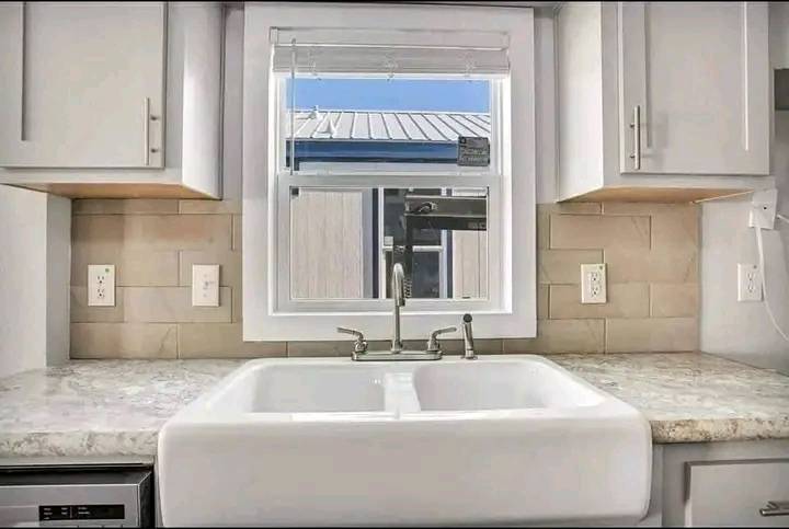 Bright kitchen with a farmhouse sink under a window. Light gray cabinets and beige backsplash create a fresh, inviting atmosphere.