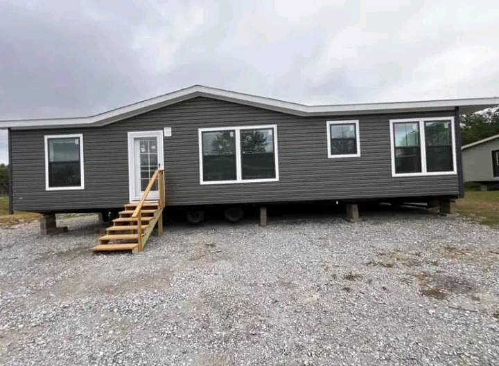 A gray modular home with a peaked roof sits on a gravel lot. It features multiple large windows and a small wooden staircase leading to a white door. Overcast sky.