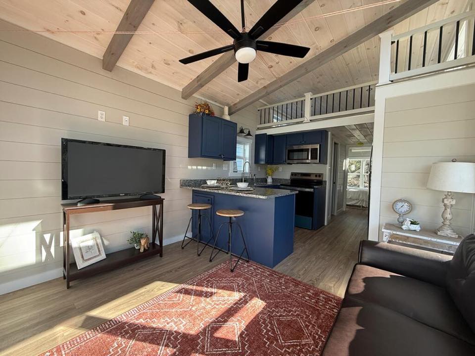 Modern tiny home interior with a cozy atmosphere. Features a blue kitchen island with stools, wood beams, flat-screen TV, ceiling fan, sofa, and red rug.