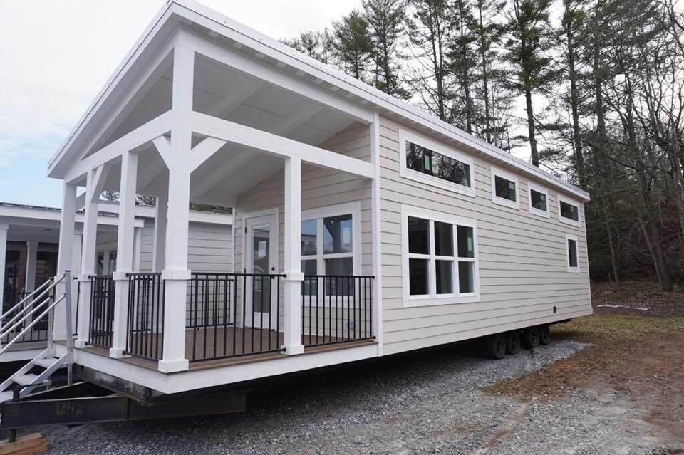 A modern tiny house on wheels with beige siding, large windows, and a covered porch with white pillars. It's set against a backdrop of tall trees.