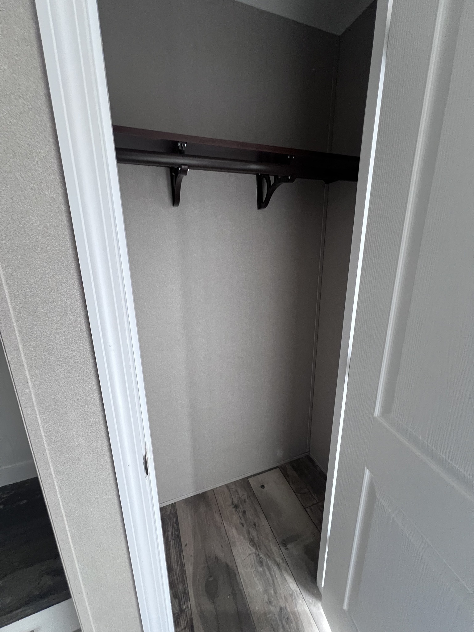 Empty closet with open white door, gray walls, and dark wooden rod. The floor is a mix of dark and light wood planks, conveying a minimalist style.