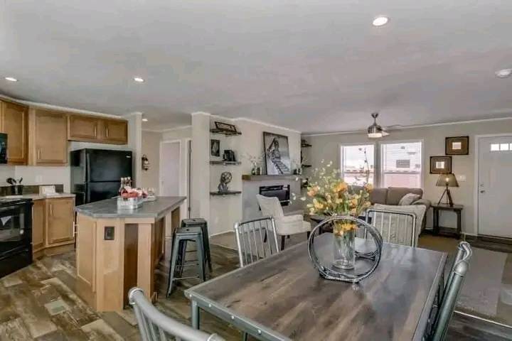 Modern open-plan kitchen and living area with wood floors. The kitchen features an island and black appliances. The living room has a fireplace, cozy seating, and wall decor. A rustic dining table with flowers adds warmth. Bright, inviting atmosphere.