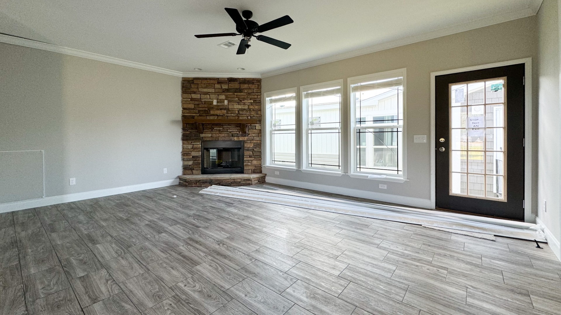 Spacious empty living room with wood-style tile flooring, a stone fireplace, ceiling fan, and large windows next to a glass-paneled door.