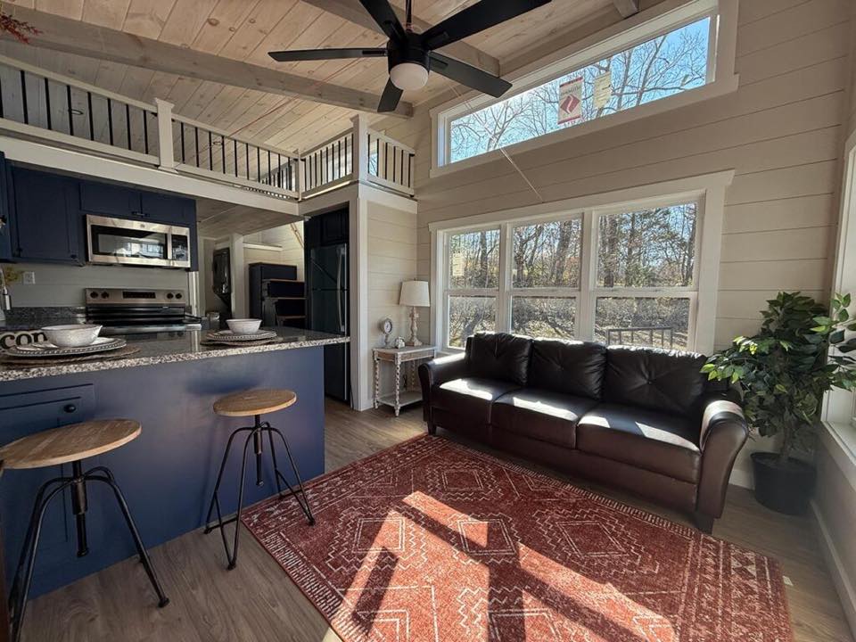Cozy living area with high ceilings and natural light. Features a brown sofa, red rug, blue kitchen island, and wooden stools, evoking warmth and comfort.