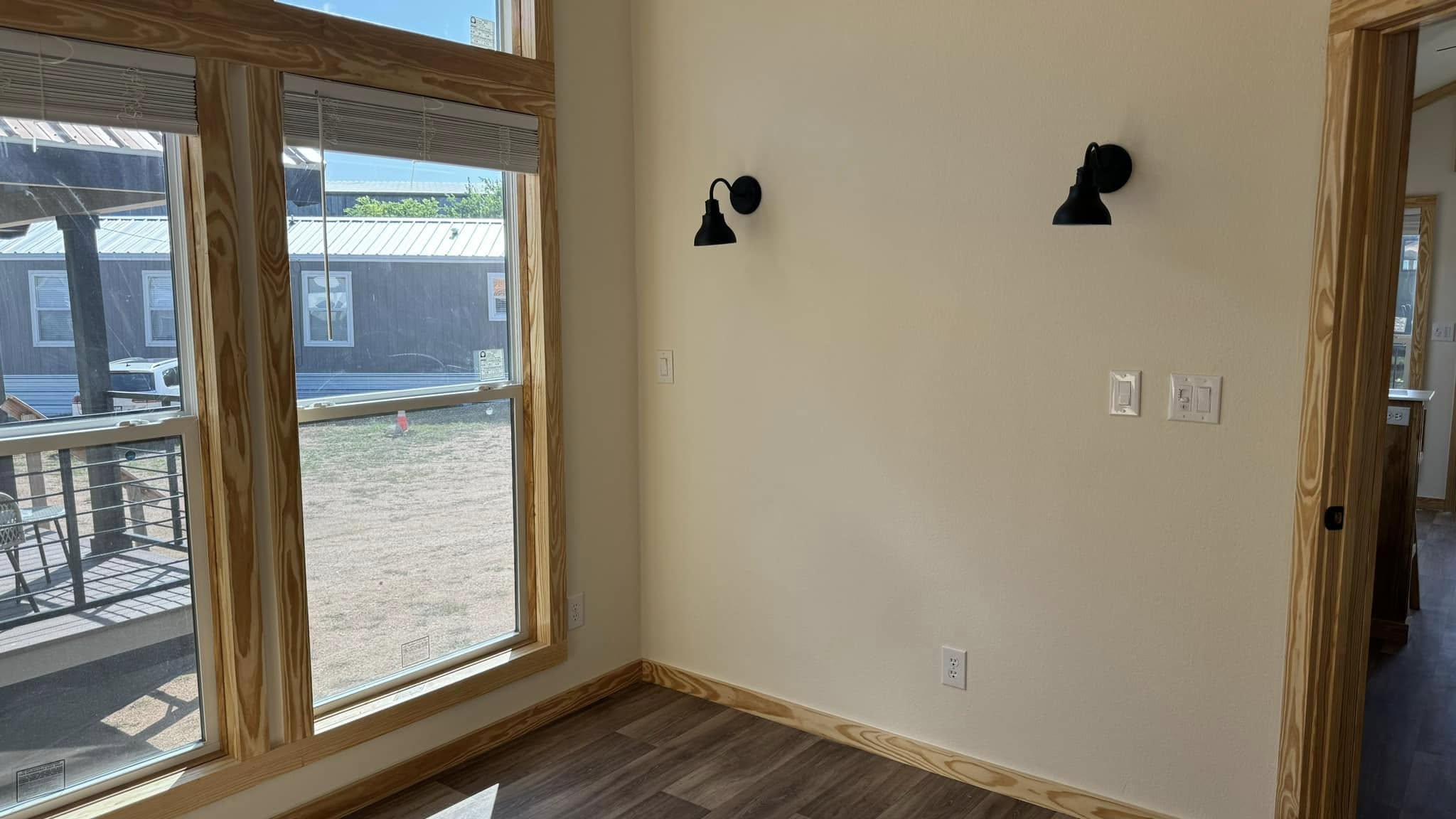 A room with large, wooden-framed windows on the left, casting natural light on wood floors. Two black wall sconces are mounted on a beige wall. Cozy and bright.