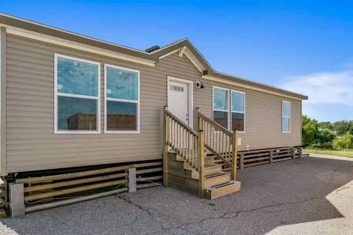 A beige mobile home with a gabled roof and front steps. Four large windows line the front, and the surroundings include a paved area and green landscape.