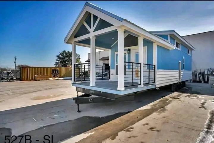 A blue and white tiny house on wheels sits on a concrete lot, showcasing a pitched roof and a covered porch. The atmosphere is bright and sunny.