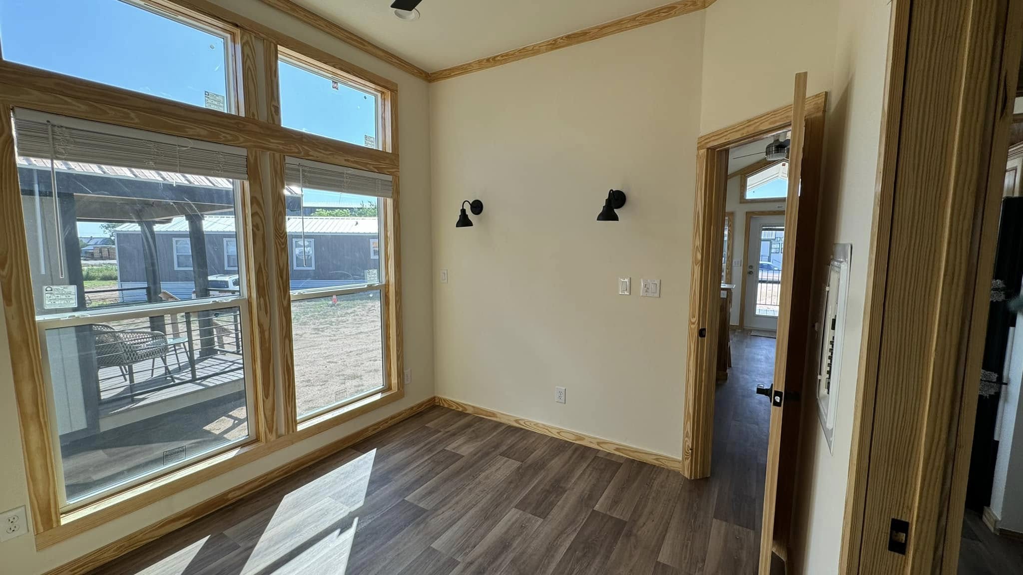 A sunlit, empty room with large windows, light wood trim, and dark wall sconces on cream walls; the door opens to an adjoining space.