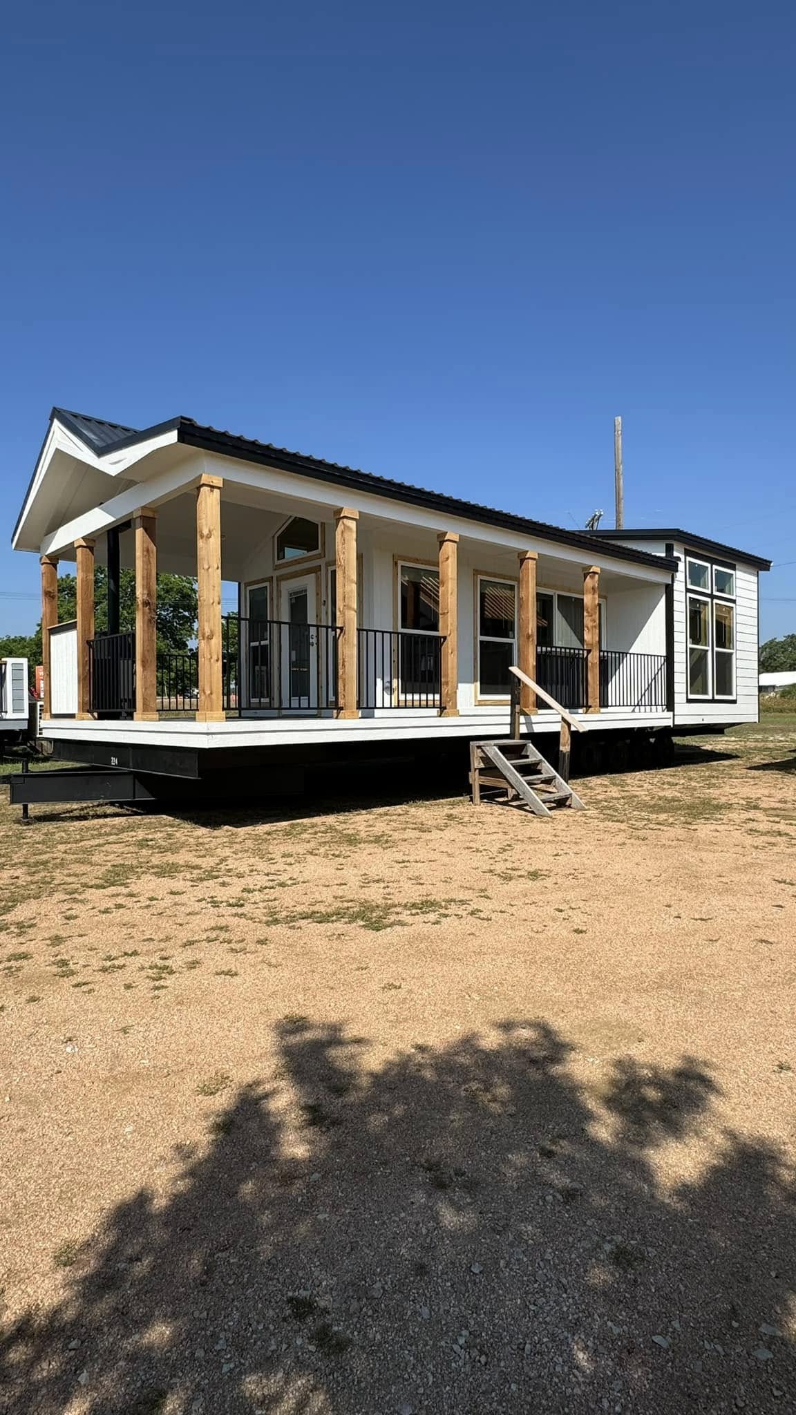 A modern tiny home with a gabled roof and wood accents sits on a gravel lot under a clear blue sky. It has a small porch and simple steps.
