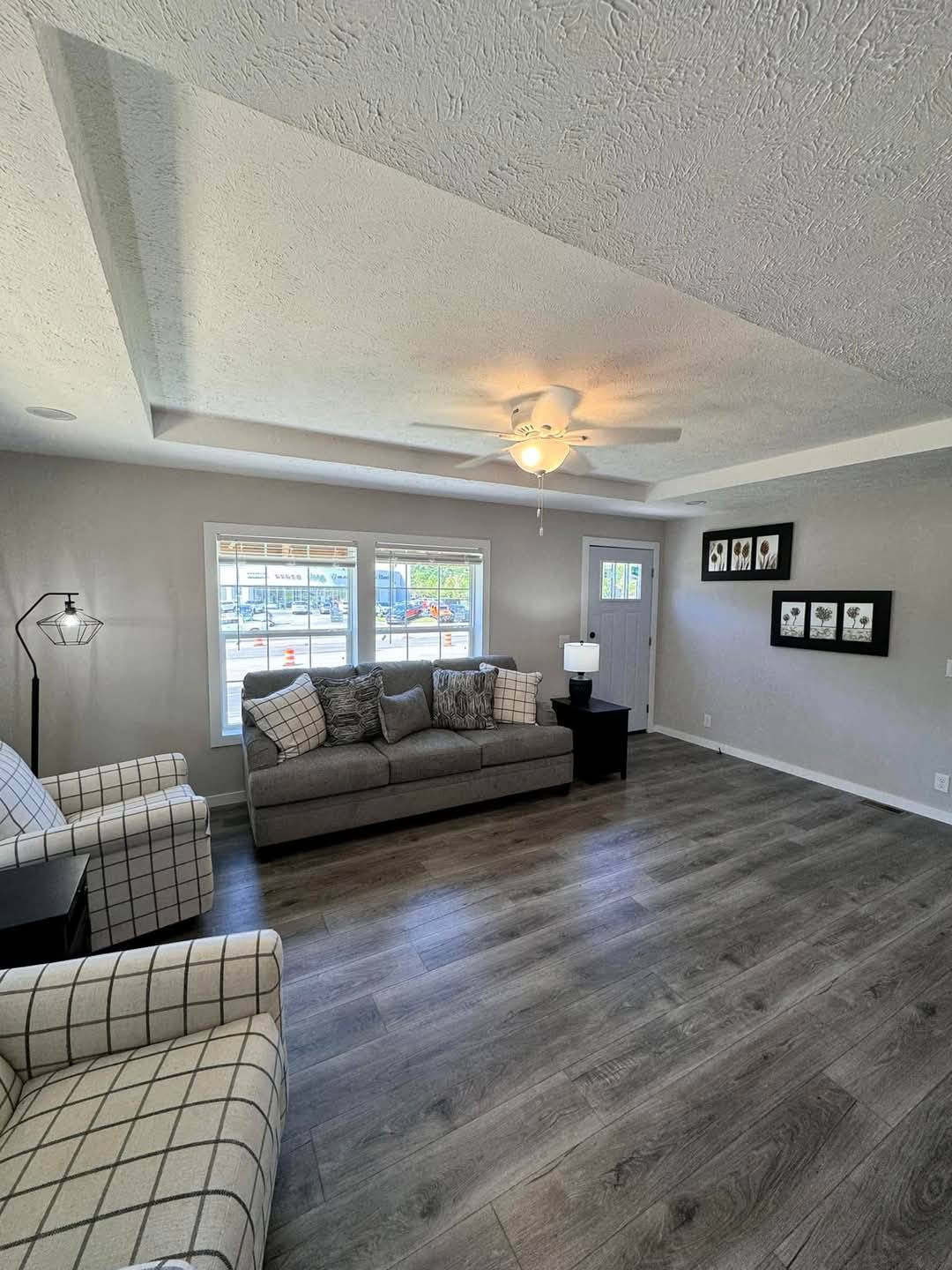 A modern living room with gray walls and wood flooring. Features a gray sofa, plaid armchairs, and framed art. Large window allows natural light.