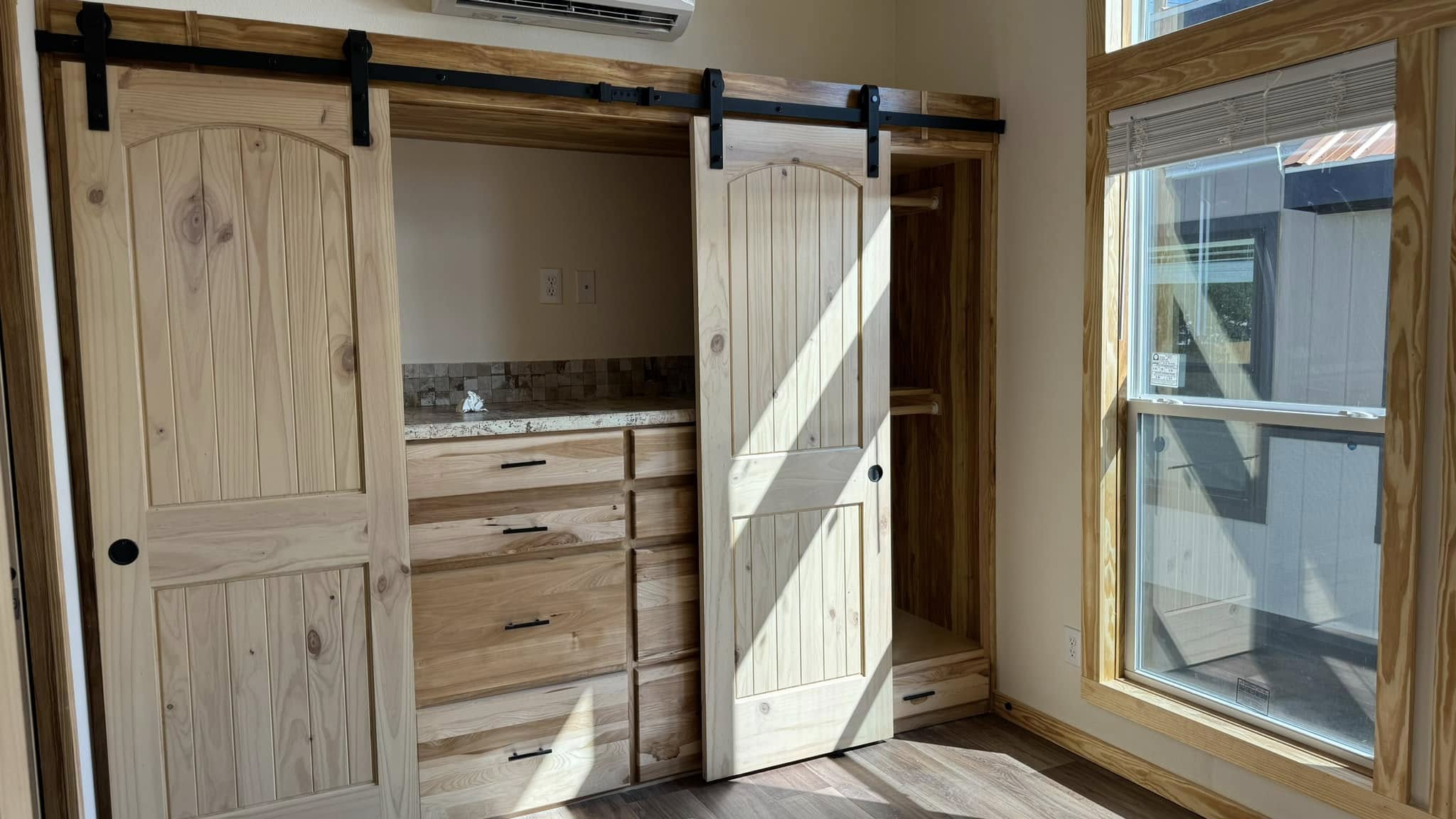 A cozy room with sliding wooden barn doors, revealing a dresser and shelves. Sunlight streams through large windows, creating a warm, inviting atmosphere.