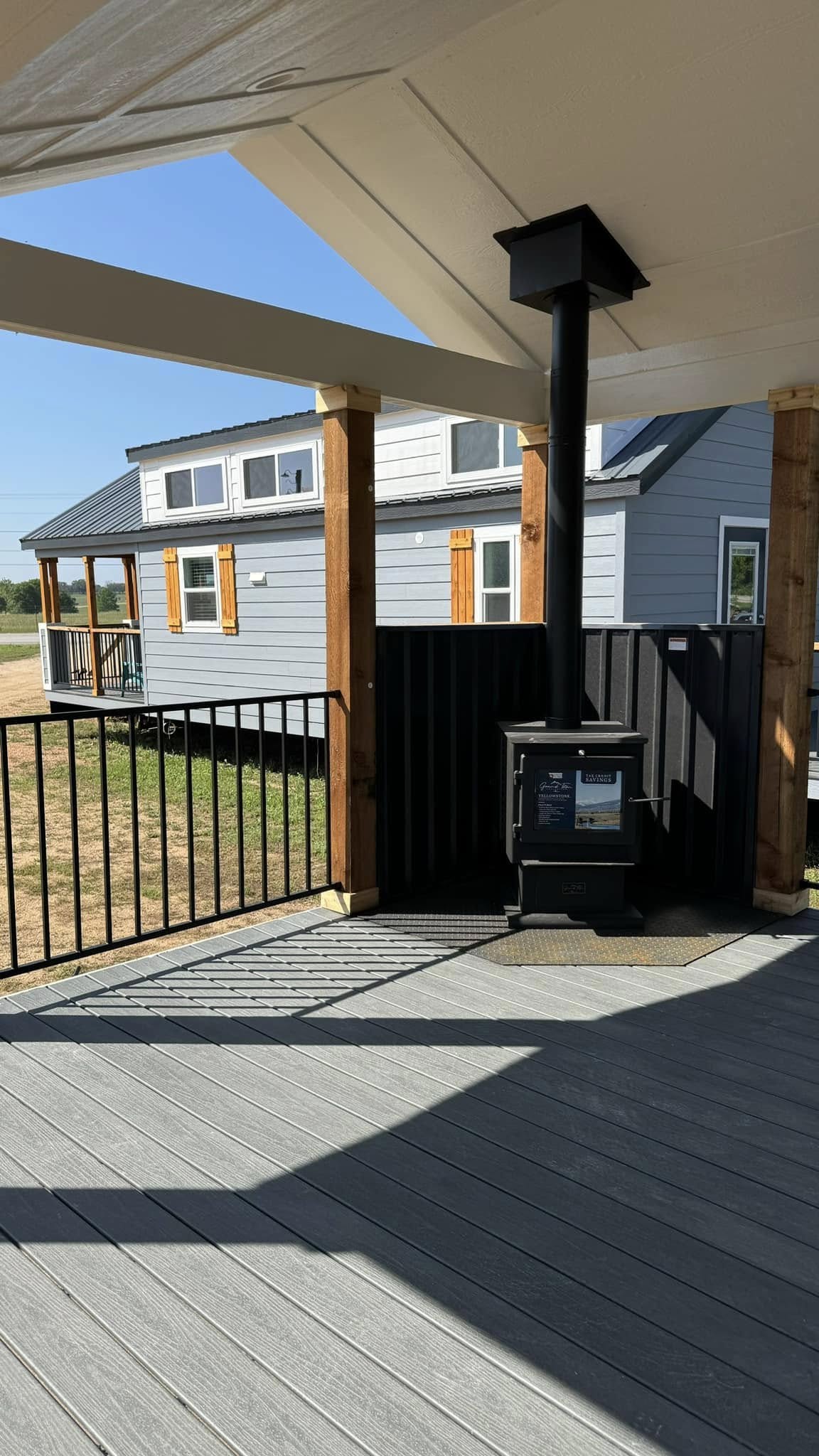 Covered patio with wooden and metal railing, featuring a small black wood stove. In the background, a modern tiny house with blue siding is visible.