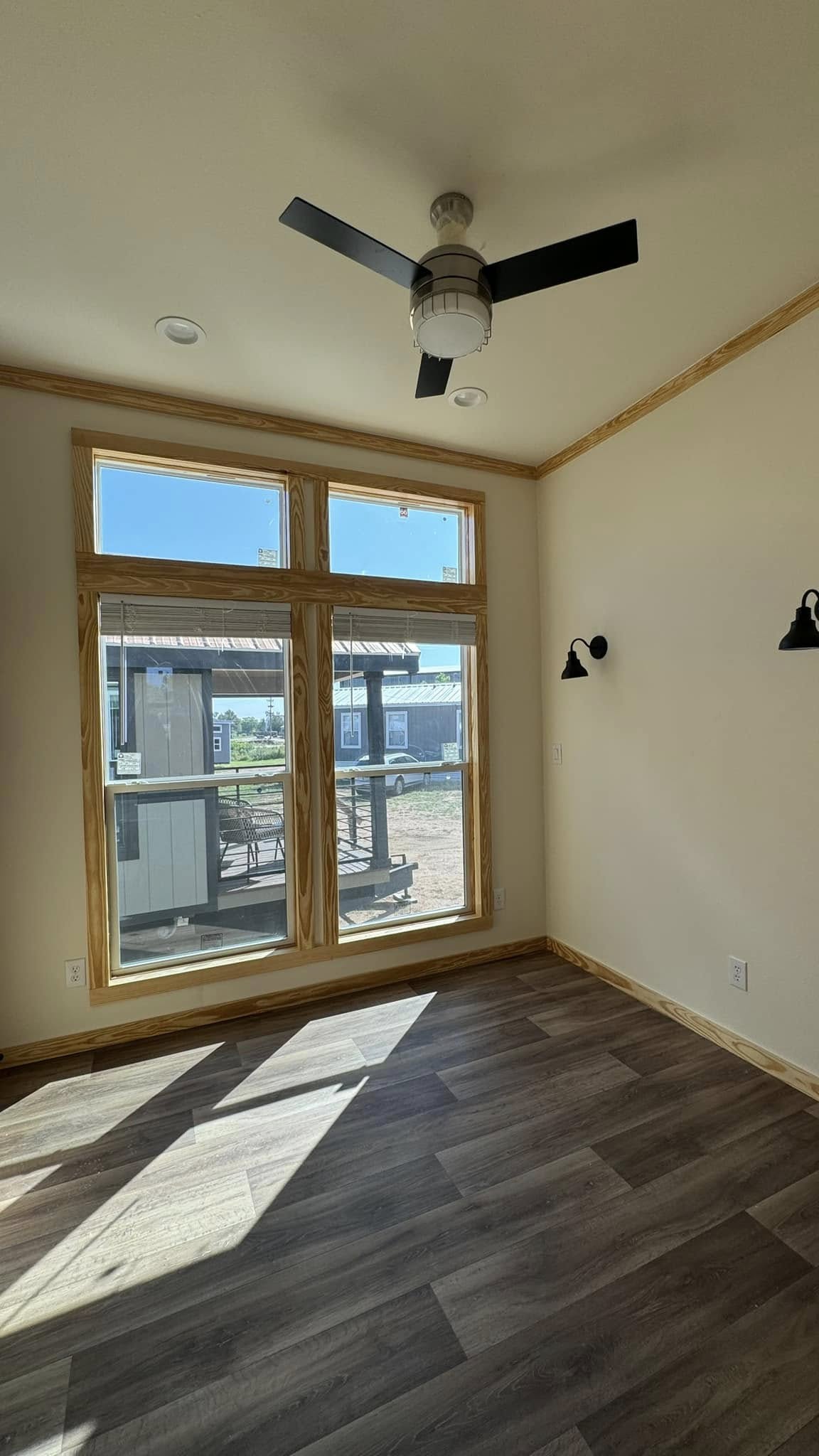 A sunlit room with wooden floors and large windows framed in light wood, showing an outdoor view. A ceiling fan and wall lamps add a modern touch.