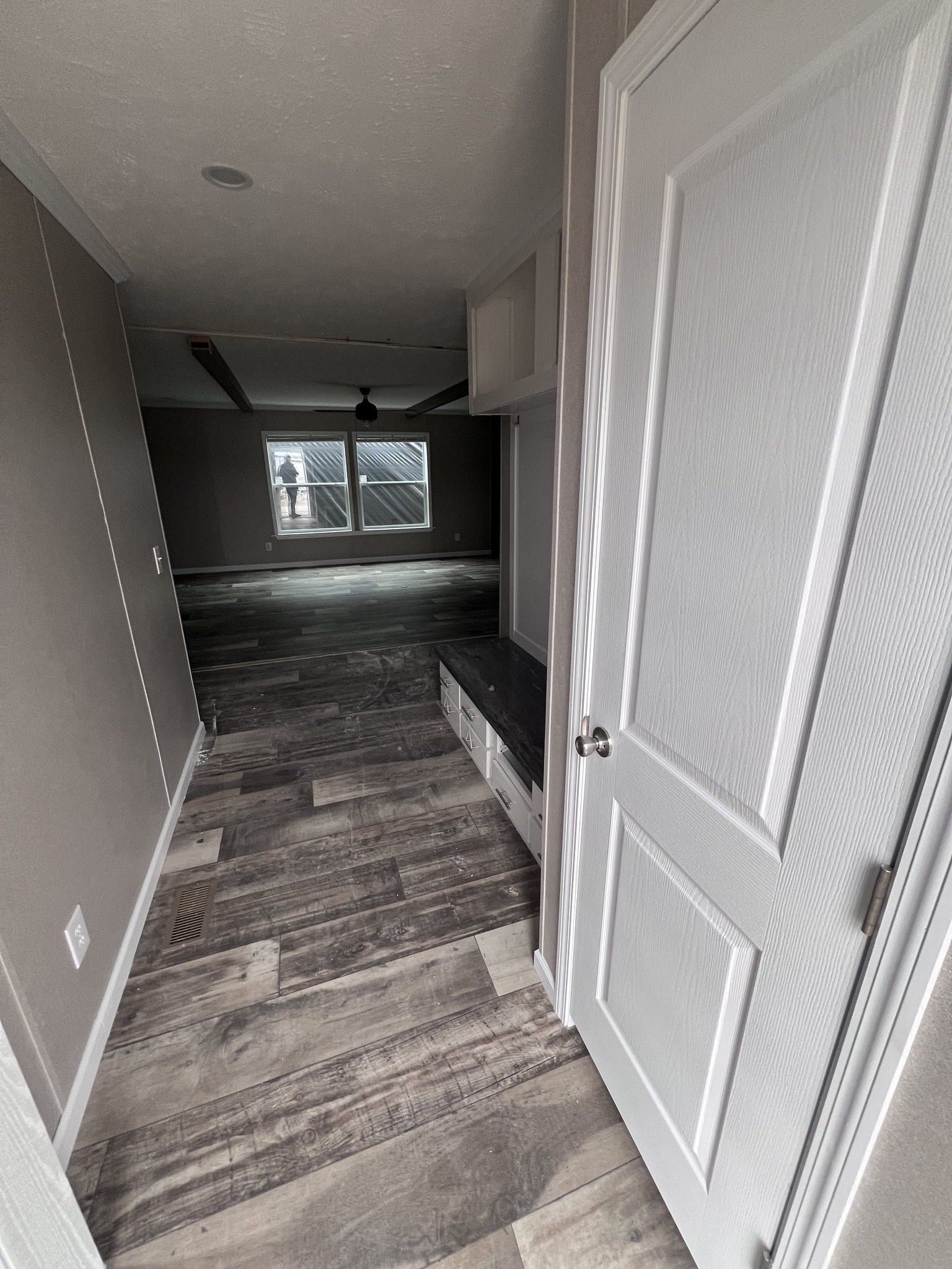 Narrow hallway with light gray wood flooring leading to a spacious living area. Large windows at the end let in natural light. Calm, modern atmosphere.