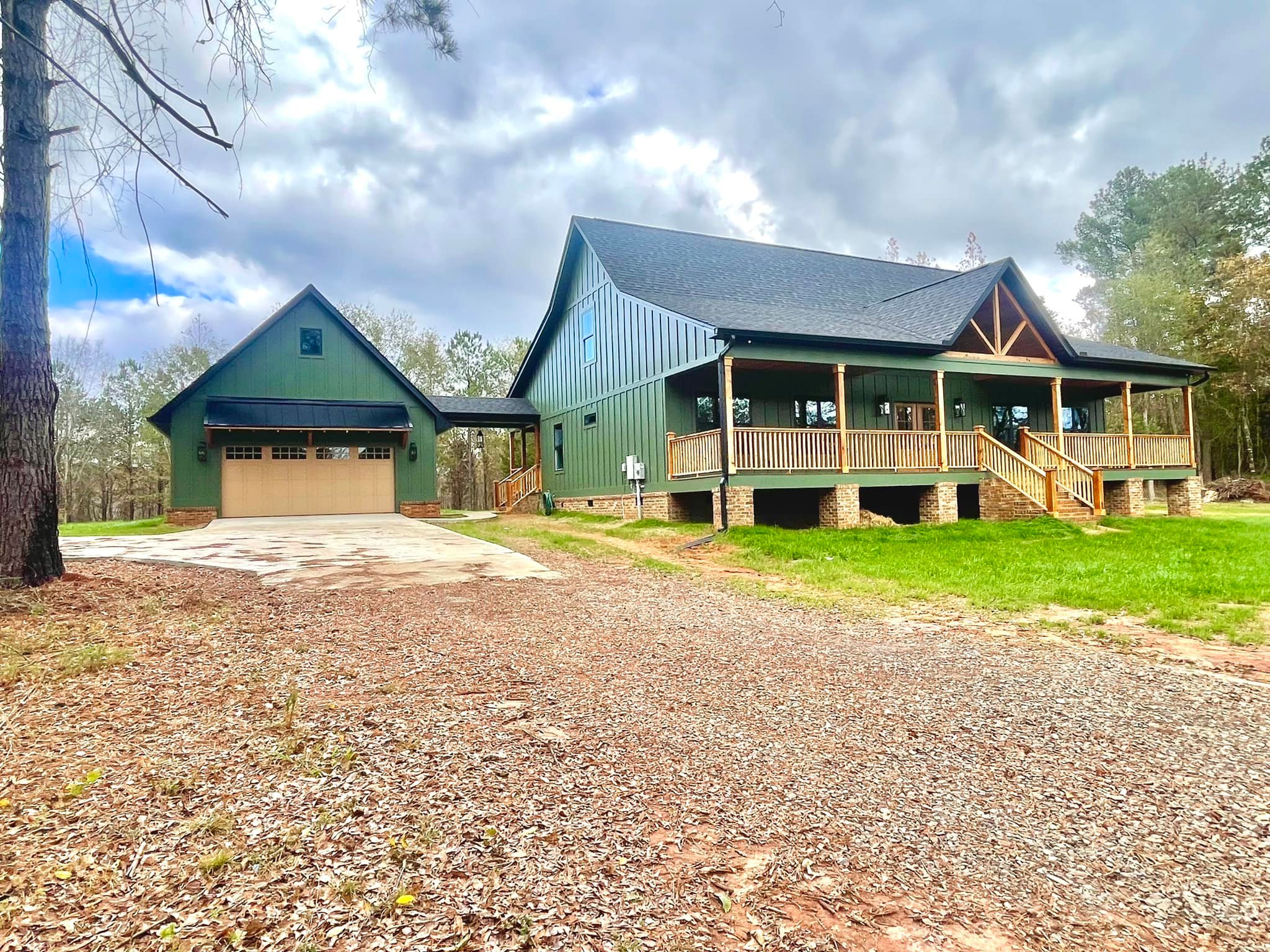 A green two-story house with a wraparound porch is surrounded by tall trees under a cloudy sky. A gravel driveway leads to an attached garage.