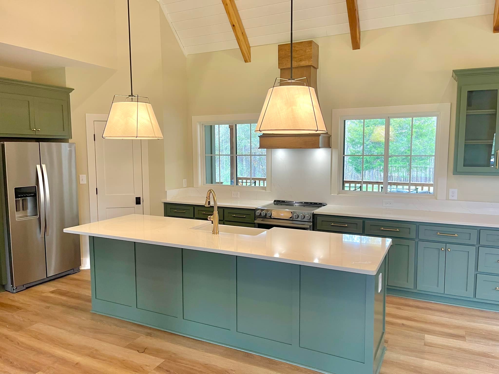 Modern kitchen with light green cabinets, white countertops, and two large pendant lights. Stainless steel fridge, wood beams, and large windows. Cozy and airy feel.