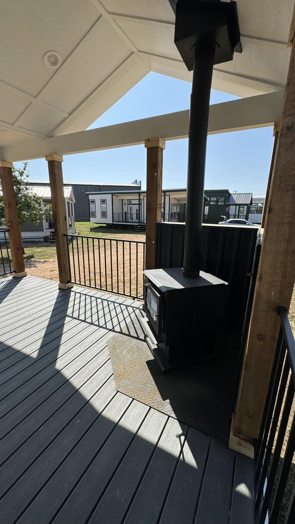 A porch with a wood-burning stove, wooden posts, and a metal railing. In the background are modern tiny houses under a clear blue sky, conveying tranquility.