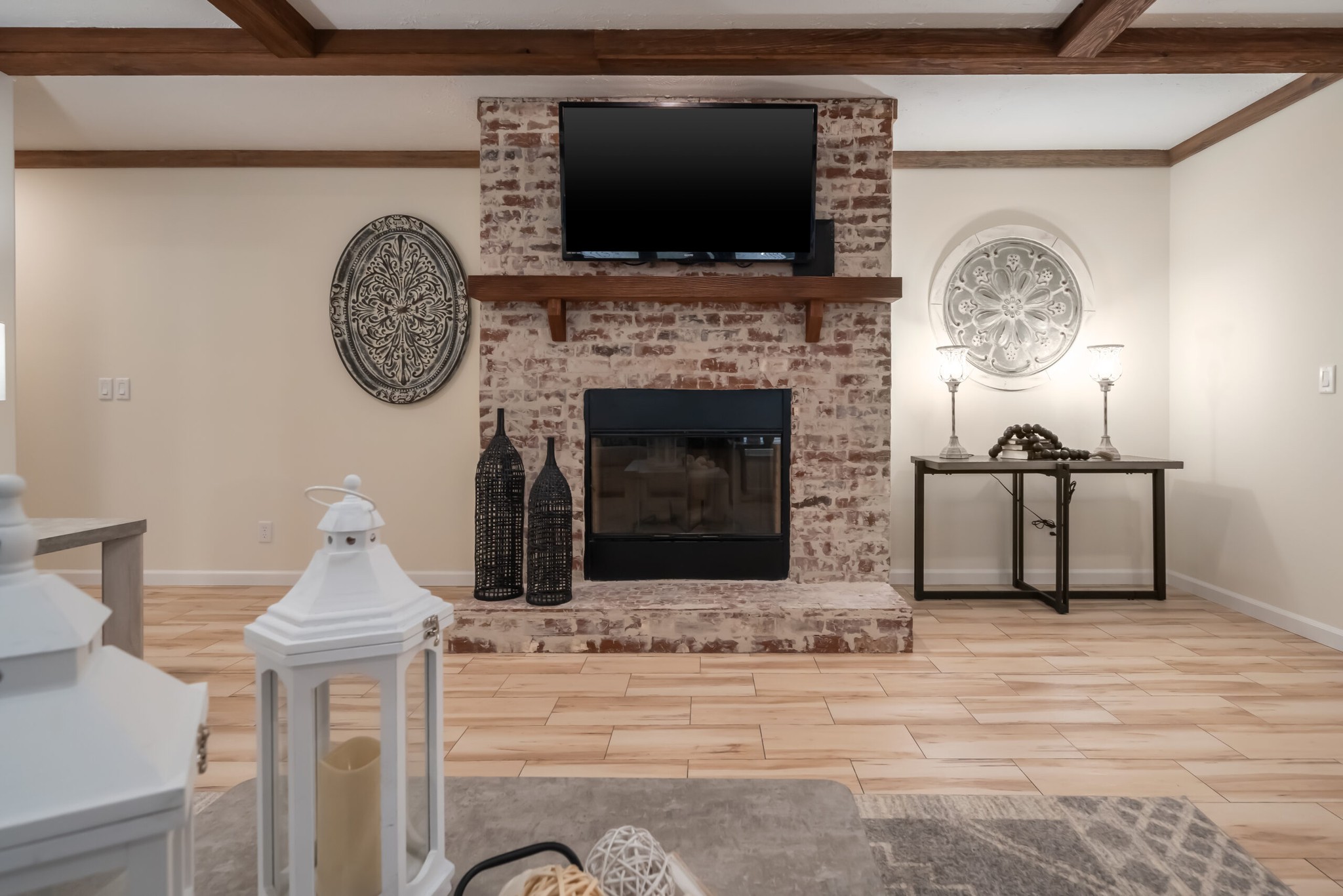 A cozy living room features a rustic brick fireplace with a mounted TV above. Decorative lanterns and wall art add charm. Neutral tones create a warm ambiance.