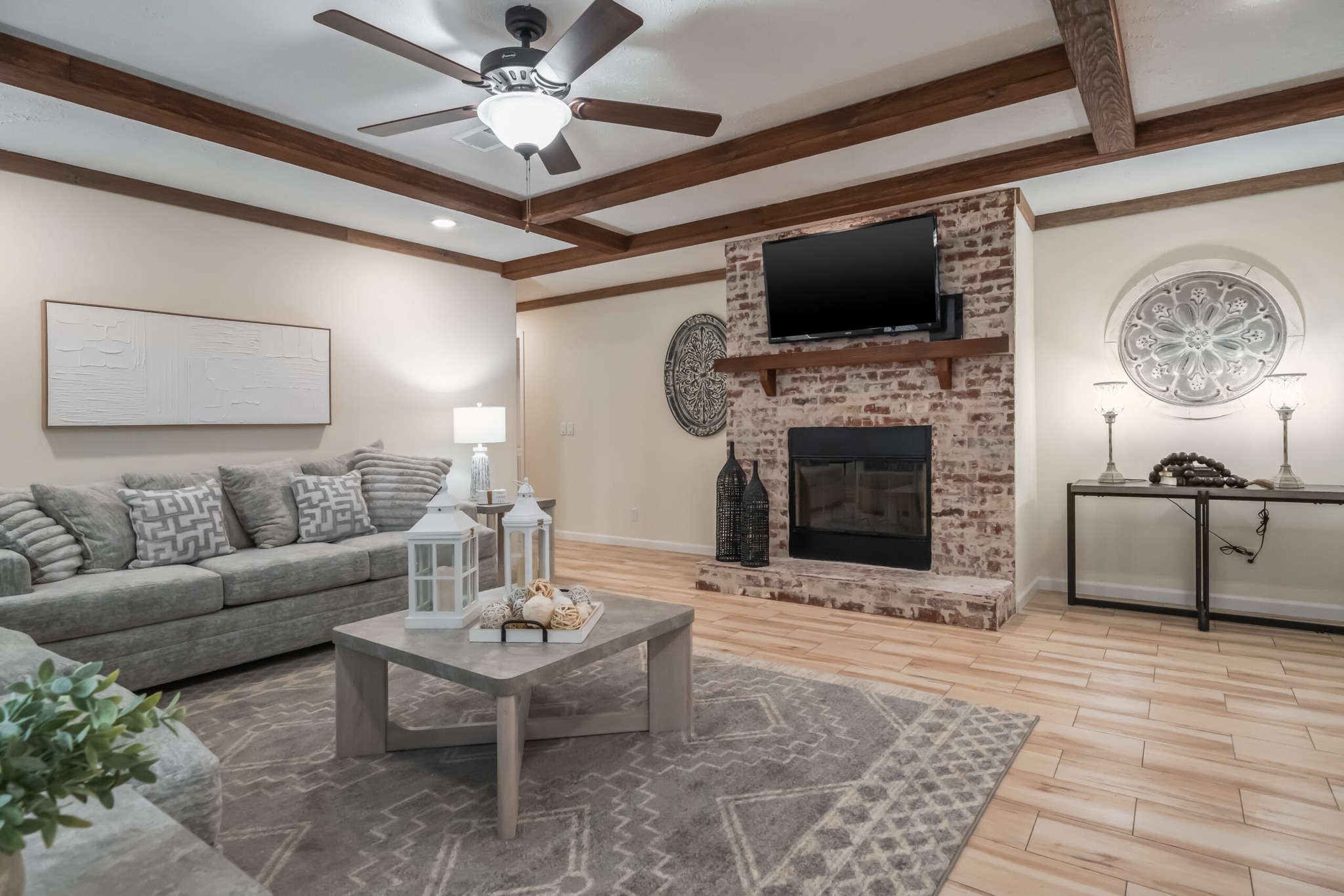 Cozy living room with beige walls, wood ceiling beams, and a brick fireplace. Gray sectional sofa, geometric rug, and rustic decor create a warm atmosphere.