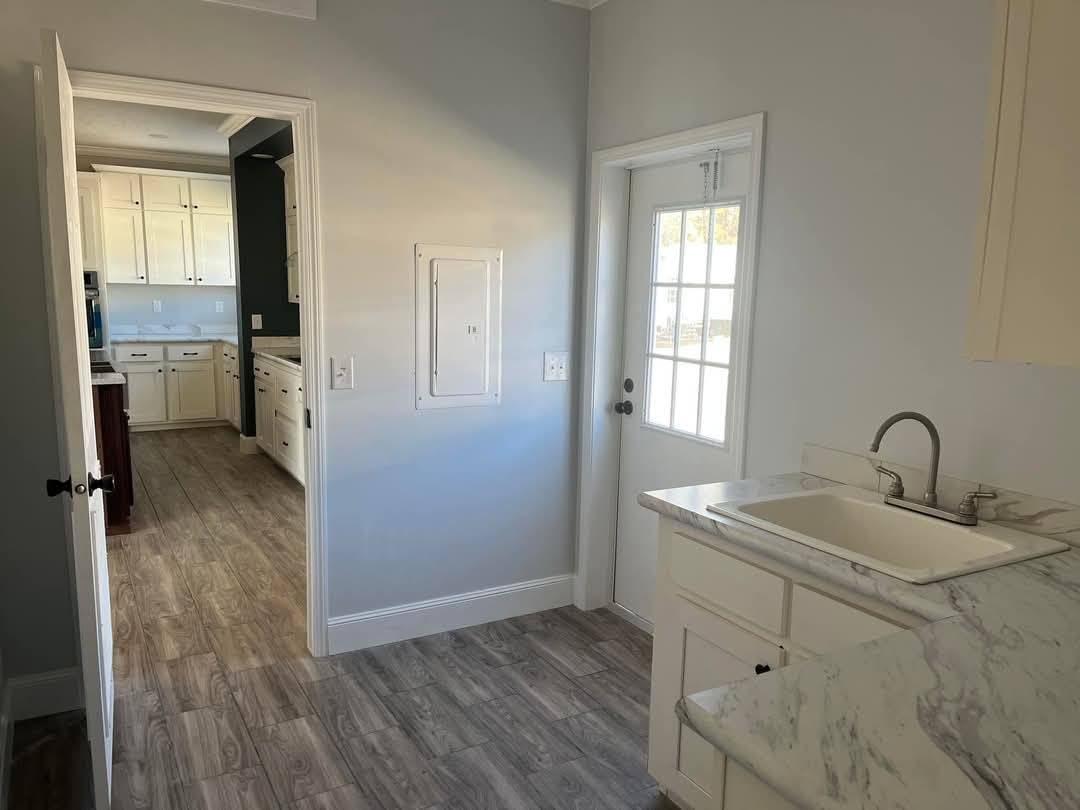 Bright kitchen with gray walls and light wood flooring. White cabinets and marble countertops surround a sink. A door with glass panes opens to the outside.