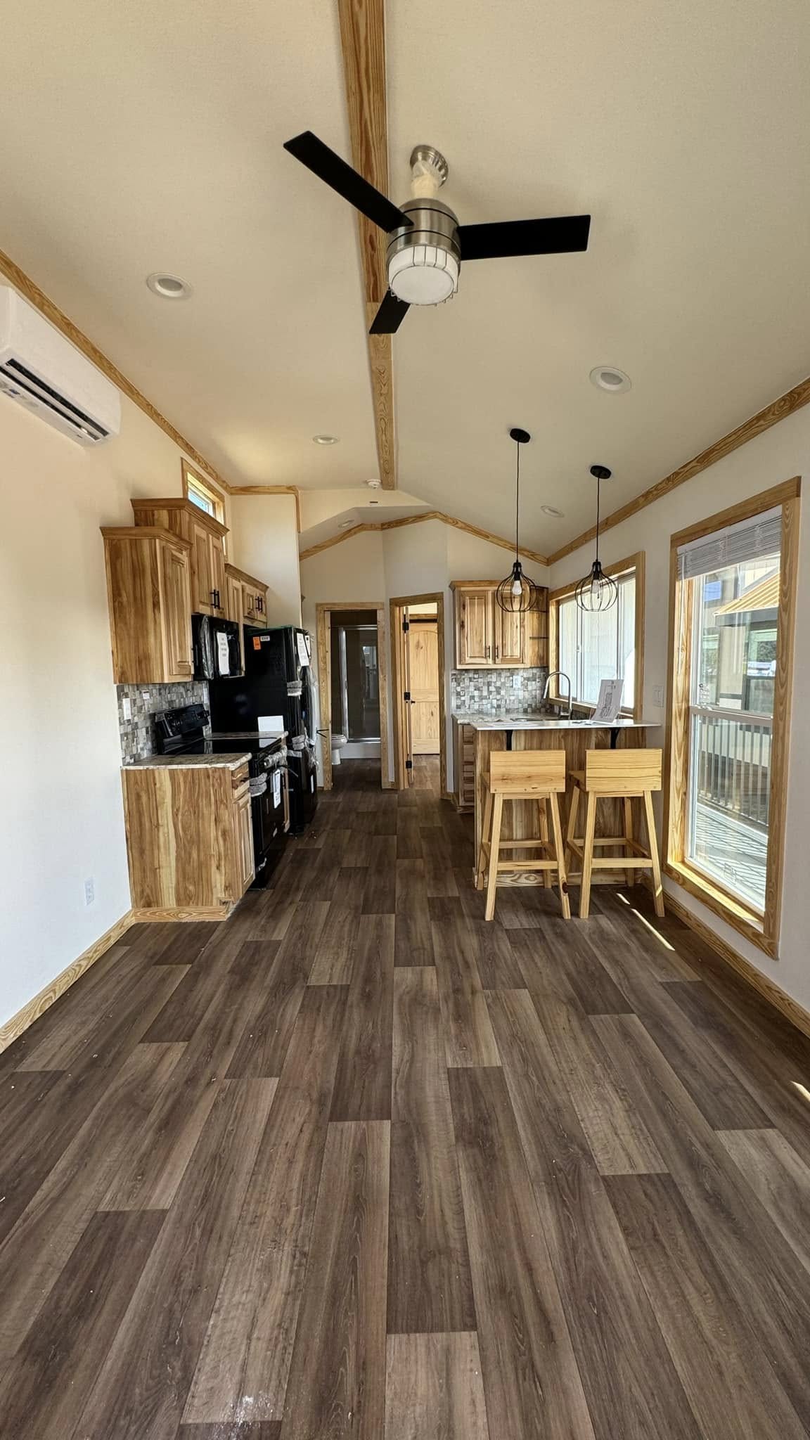 Modern tiny home interior with a high ceiling, wood accents, and large windows. Features a compact kitchen, bar stools, and a ceiling fan, creating an open, airy feel.