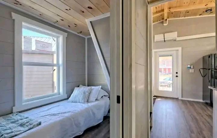 Cozy interior of a tiny house featuring a single bed with striped pillows by a window. Light wood flooring leads to a bright entryway, creating an inviting and compact living space.