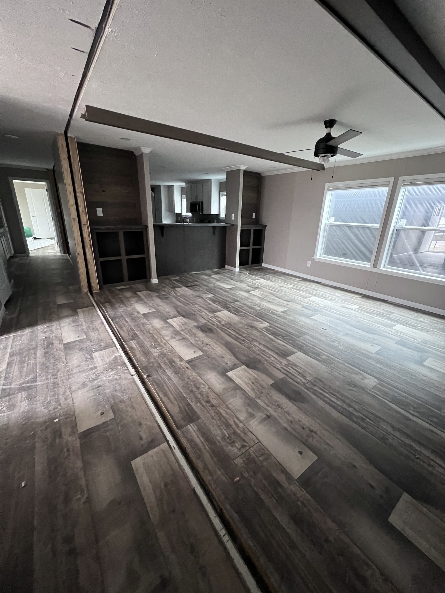 Spacious room with wooden flooring, a ceiling fan, and large windows letting in natural light. The open-plan area leads to a kitchen with dark cabinets.