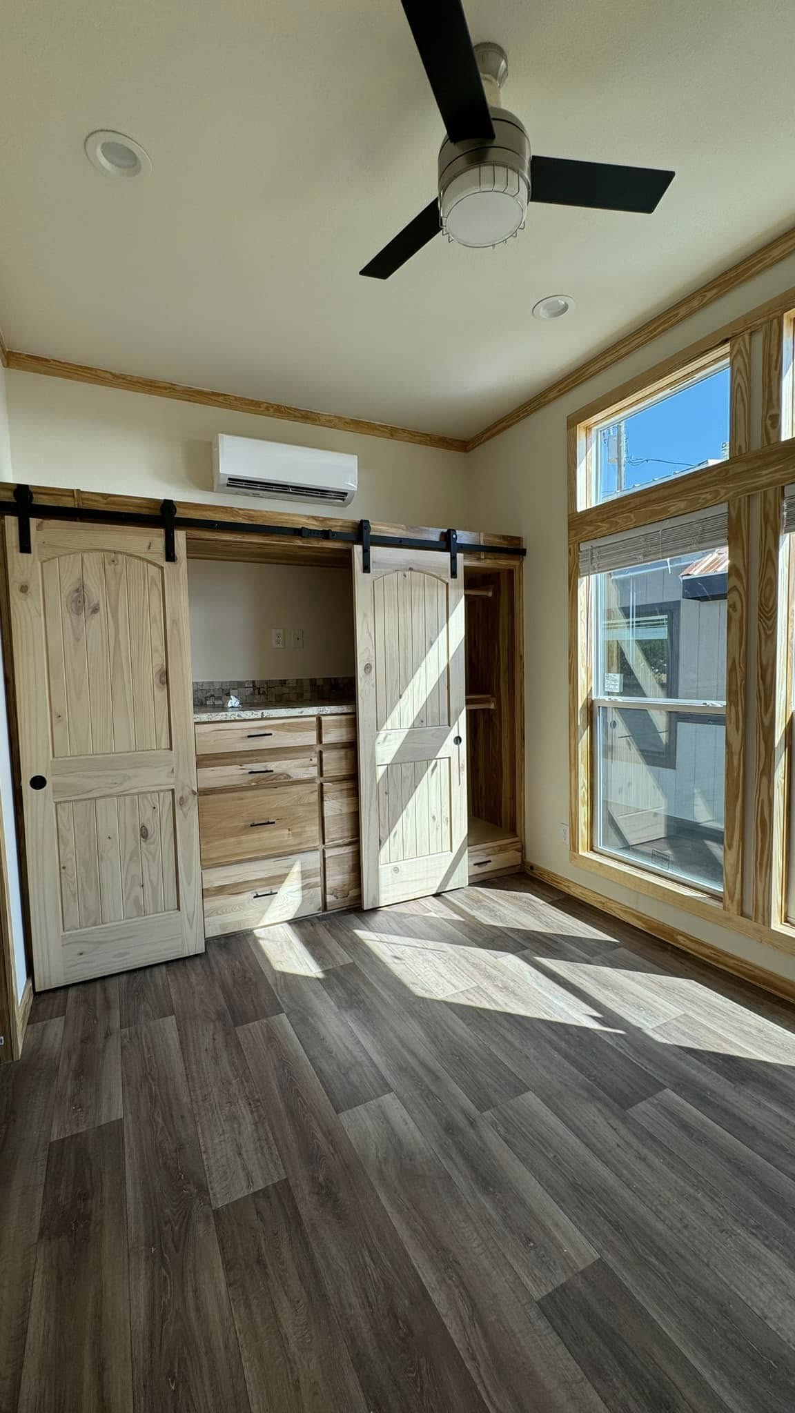 Sunlit room with dark wood flooring, sliding barn doors, and large windows. A white ceiling fan hangs above, creating a cozy, rustic atmosphere.