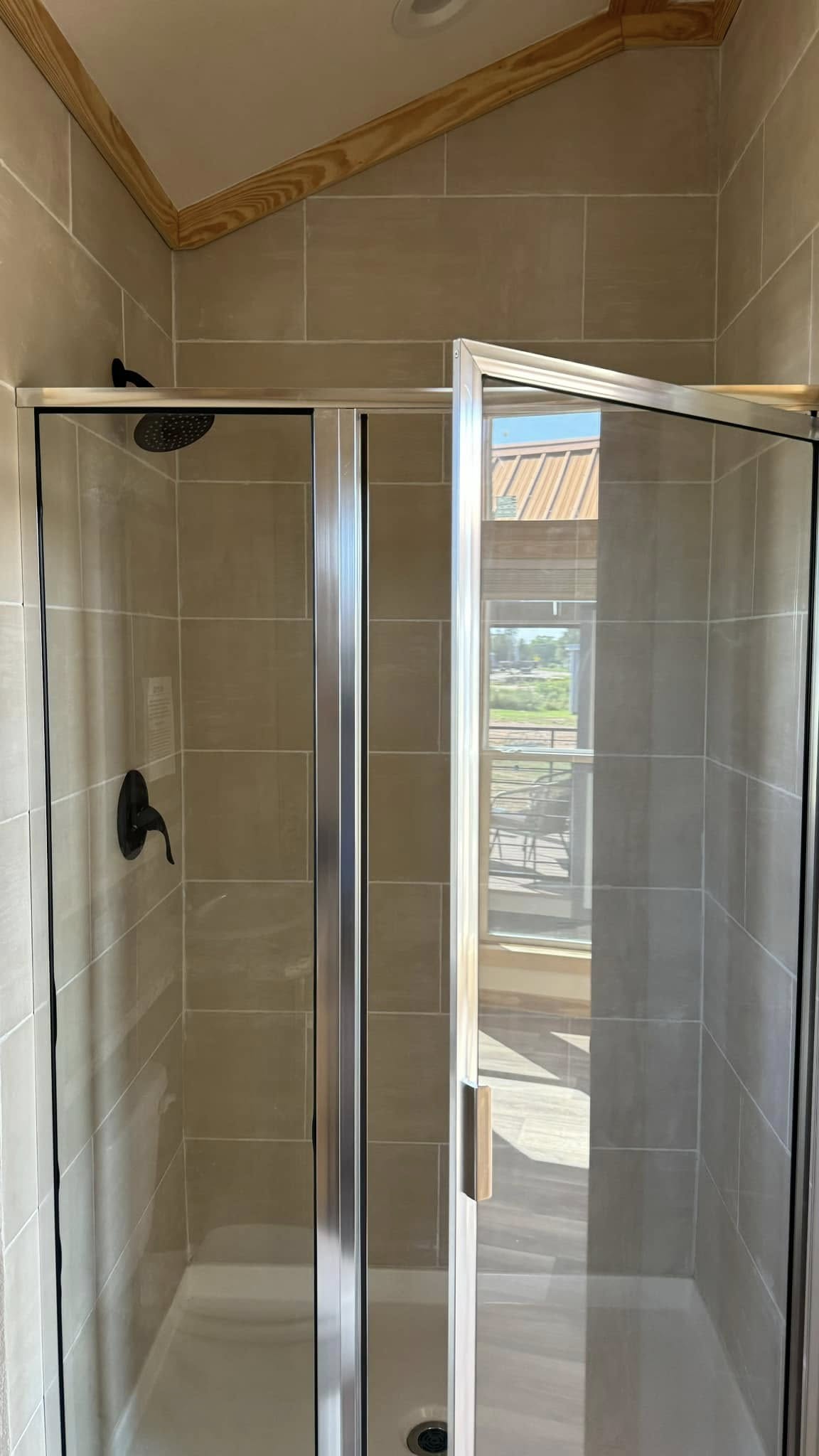 Compact bathroom with a beige-tiled shower, sliding glass doors, dark fixtures, and a wooden ceiling trim. Sunlight streams in, creating a serene atmosphere.